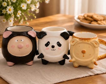 May include: Three ceramic mugs shaped like animals. The left mug is black with pink ears and cheeks. The center mug is white with black panda features. The right mug is light brown with small ears and a simple nose, all featuring handles.
