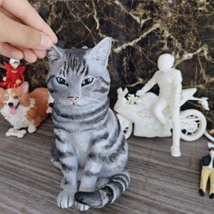 May include: A gray and black tabby cat figurine with blue eyes, sitting in an upright position. The cat has distinct dark stripes across its head, back, and tail, with a light gray chest and white whiskers. The figurine is sculpted with realistic fur texture.