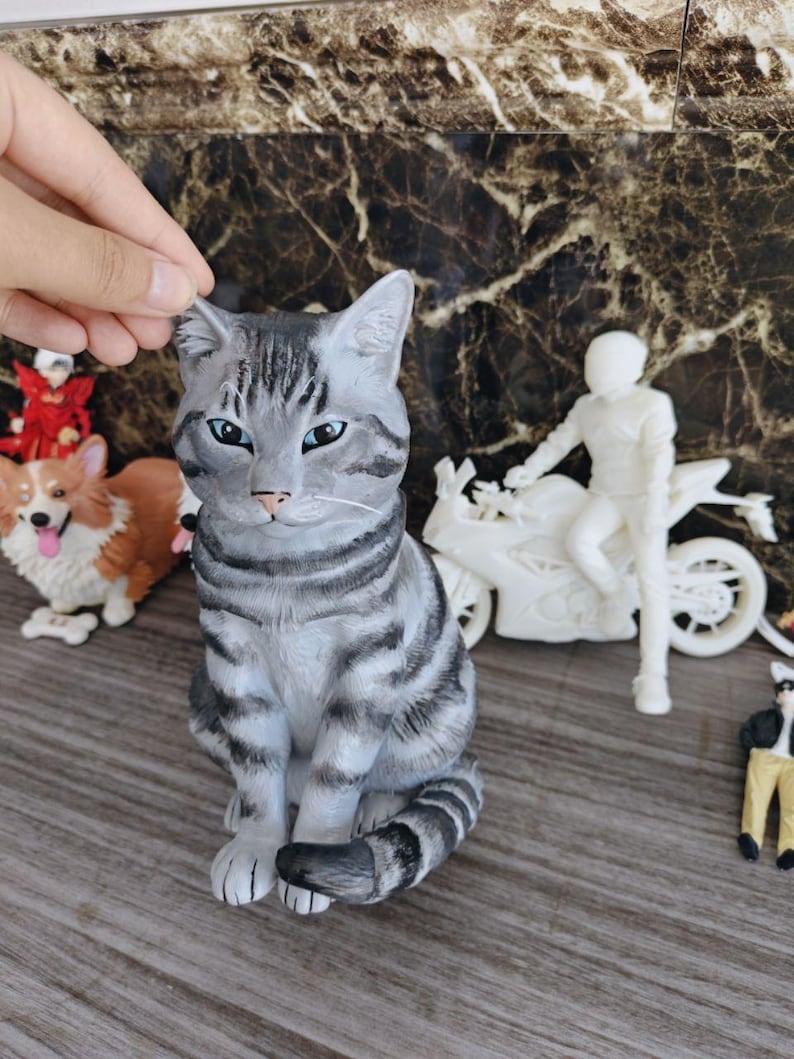 May include: A gray and black tabby cat figurine with blue eyes, sitting in an upright position. The cat has distinct dark stripes across its head, back, and tail, with a light gray chest and white whiskers. The figurine is sculpted with realistic fur texture.