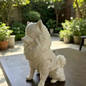 May include: A cream-colored figurine of a Pomeranian dog in a seated position. The dog has a thick, textured coat, pointed ears, and is wearing a simple, smooth-textured vest. The sculpture is rendered in a solid, matte, off-white material.