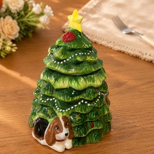 Op de afbeelding: Een keramische pot in de vorm van een kerstboom met een glanzende groene afwerking, voorzien van wit geschilderde slingers, kleine rode en gele ornamenten en een gele ster als piek. Een bruin-witte beagle is onderaan de boom gebeeldhouwd.
