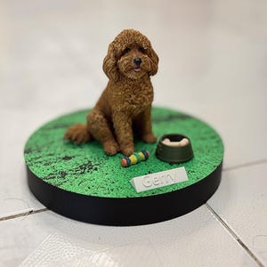 May include: A brown curly-haired dog figurine sits on a circular green base with a textured surface. Beside the dog is a small green bowl containing a bone and a colorful toy. A white rectangular nameplate with the text Gemy is attached.