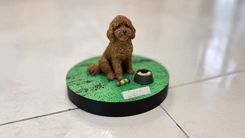 May include: A brown curly-haired dog figurine sits on a circular green base with a textured surface. Beside the dog is a small green bowl containing a bone and a colorful toy. A white rectangular nameplate with the text Gemy is attached.