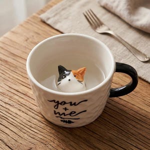 Op de afbeelding: Witte keramische mok met een zwarte handgreep en een getextureerde buitenkant. De mok heeft een driedimensionaal kattengezicht aan de binnenkant en de woorden "you + me" in zwart schrift op de zijkant. Een vork ligt op een linnen servet.