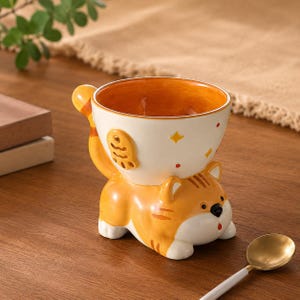 Op de afbeelding: Een keramische kom met een oranje binnenkant, ondersteund door een cartoon kattenfiguur. De kom is wit met oranje en gele accenten, waaronder een visontwerp. De kat is oranje en wit met zwarte details. Een gouden lepel ligt in de buurt.