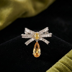 May include: A silver bow brooch featuring a central yellow gemstone and a dangling, teardrop-shaped yellow gem. The bow is adorned with rectangular, clear crystals. The brooch is set against a black backdrop.