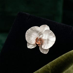 May include: A silver orchid flower brooch with a copper-coloured centre. The brooch is set against a black background with a green velvet accent.