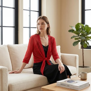 Peut inclure: Boléro rouge à manches trois-quarts, noué à la taille, porté sur une robe noire. La personne est assise sur un canapé beige dans une pièce lumineuse, avec une plante et une table basse.