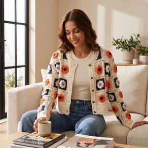 Könnte beinhalten: Cremefarbene Häkeljacke mit quadratischen Blumenmustern in Orange, Lila und Weiß. Die Strickjacke mit Knopfleiste wird mit einem weißen Oberteil und einer blauen Jeans getragen. Eine Kaffeetasse und Bücher liegen auf einem Holztisch.