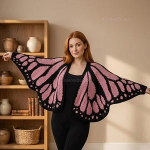 Puede incluir: Un cárdigan negro y rosa con forma de ala de mariposa. El cárdigan presenta un borde negro con patrones de alas rosas. La mujer de la imagen lleva un atuendo negro. El fondo incluye una estantería de madera con cerámica y libros.