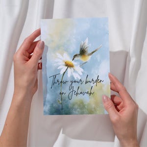 Puede incluir: Tarjeta de felicitación con una pintura de acuarela de un pájaro y una margarita. La tarjeta tiene un fondo azul y amarillo suave con el texto "Throw your burden on Jehovah" en escritura elegante.