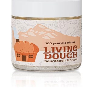 May include: A clear glass jar with a white lid, labeled "100 year old Alaska Living Dough Sourdough Starters." The label features a mountain range, a bread house, and various bread illustrations. The jar contains a light-colored powder.