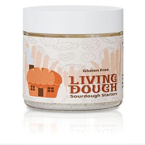 以下が含まれることがあります： 白い蓋が付いた透明なガラス瓶で、「Gluten Free Living Dough Sourdough Starters」とラベルされています。ラベルには、漫画のパンの家とパンのイラストが描かれています。瓶には粒状の物質が入っています。