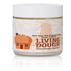 Puede incluir: Un tarro de vidrio transparente con tapa blanca, etiquetado "Living Dough Sourdough Starters". La etiqueta presenta una ilustración de dibujos animados de una casa de pan y el texto "900 Year Old Organic Fresh Yeast Culture."