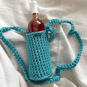 May include: A turquoise crochet wine bottle holder with a strap. The holder is designed to carry a wine bottle, visible inside. The background is a white, textured fabric.