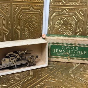 May include: A Singer Hemstitcher attachment for family sewing machines. The attachment is in a cardboard box, with the box open. The box has a green and white label. The attachment is silver and has several moving parts.