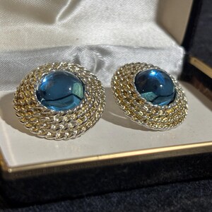 May include: A pair of round clip-on earrings featuring a central dark blue cabochon stone surrounded by a textured, braided gold and silver tone metal border. The earrings are presented inside a black jewelry box with a white satin lining.