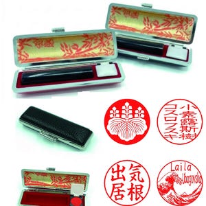 Op de afbeelding: Een verzameling Japanse hanko-stempels en -etuis. De open etuis tonen een zwarte cilindrische stempel, een wit inktkussen en een rood inktkussen. De etuis hebben een zilveren frame en een decoratief interieur. Er worden ook verschillende rode ronde stempels getoond.