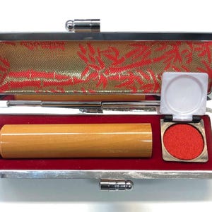 May include: Open Japanese stamp case with a wooden stamp, red ink pad, and a small white lid. The case has a silver frame and a red velvet interior. The lid is lined with gold fabric with red floral patterns.