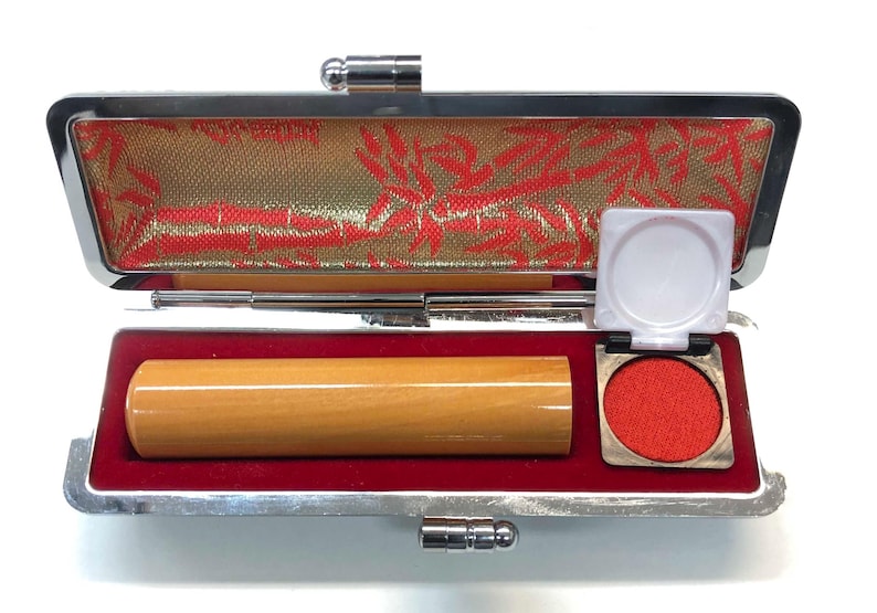 May include: Open Japanese stamp case with a wooden stamp, red ink pad, and a small white lid. The case has a silver frame and a red velvet interior. The lid is lined with gold fabric with red floral patterns.