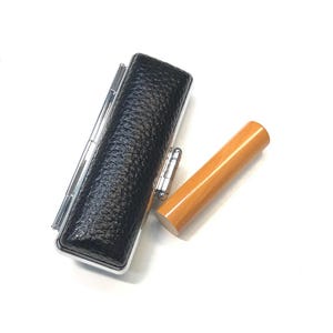 May include: A black leather stamp case with silver accents and a light brown wooden stamp. The stamp case has a textured surface. The wooden stamp is cylindrical.