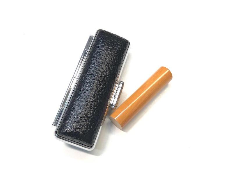 May include: A black leather stamp case with silver accents and a light brown wooden stamp. The stamp case has a textured surface. The wooden stamp is cylindrical.