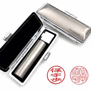 May include: A rectangular metallic case with the word TITANIUM printed on the lid, shown both closed and open. Inside the open case is a cylindrical metallic seal. Two circular red ink stamps with Chinese characters are positioned below the case.
