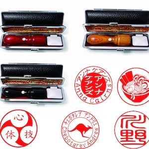 May include: A collection of wooden and black stamps in open black cases, alongside red ink stamps with Japanese text and illustrations. The stamps include a kangaroo, ramen, and other circular designs. The cases have a silver trim and a white ink pad.