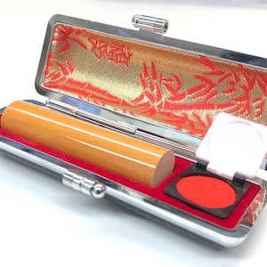 Puede incluir: Un estuche rectangular plateado para sellos con un sello de madera y una almohadilla de tinta roja. El interior del estuche está forrado con tela roja y una tela dorada estampada. El sello es cilíndrico y marrón claro.