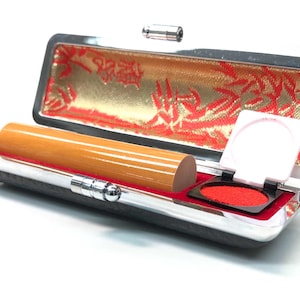 May include: A wooden stamp and ink pad set in a silver and black case. The case is open, revealing a cylindrical wooden stamp, a red ink pad, and a decorative gold and red interior lining. The ink pad is open, ready for use.