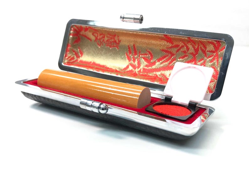May include: A wooden stamp and ink pad set in a silver and black case. The case is open, revealing a cylindrical wooden stamp, a red ink pad, and a decorative gold and red interior lining. The ink pad is open, ready for use.