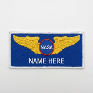 May include: A blue rectangular patch with a white border. The patch features gold embroidered wings, a red circle with "NASA" inside, and the text "NAME HERE" in white.