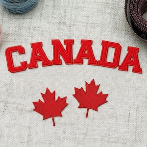 May include: Red embroidered patch with the word "CANADA" in bold, arched letters. Two red maple leaf patches are below the word. The patches are likely for sewing or ironing onto fabric.