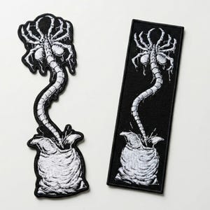 May include: Two black and white embroidered patches. One is a die-cut patch, and the other is rectangular. Both feature a detailed illustration of a facehugger emerging from an egg. The patches are likely for clothing or accessories.