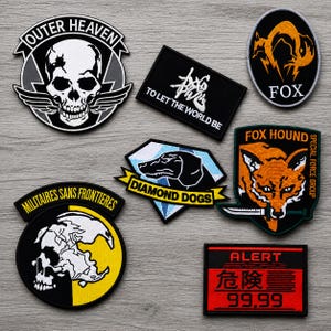May include: A collection of seven embroidered patches featuring various designs including a skull with wings labeled Outer Heaven, a rectangular patch with text To Let The World Be, a Fox logo, a Diamond Dogs patch, a Fox Hound shield, a skull patch, and an Alert sign.
