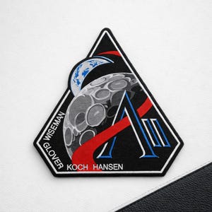 May include: A black, white, and red embroidered patch with a space-themed design. The patch features a stylized moon, Earth, and the letter "A". The names "WISEMAN", "GLOVER", "KOCH", and "HANSEN" are also visible.