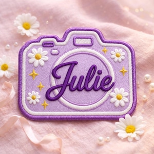 May include: A purple embroidered patch shaped like a camera, featuring the name Julie in dark purple script across the center. The design is decorated with white daisy flowers with yellow centers and small yellow four-pointed star accents.