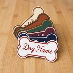 May include: A stack of bone-shaped dog tags in various colors, including white, navy, burgundy, green, and tan. The front tag reads "Dog Name" in a cursive font with a red border. The tags are stacked on a wooden surface.