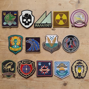 May include: A collection of embroidered patches with various designs, including a skull, a stylized eagle, and geometric shapes. The patches feature a range of colors, such as green, blue, and yellow, and are arranged on a wooden surface.