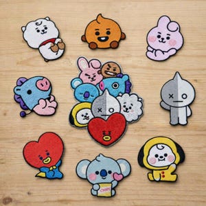 May include: A collection of embroidered patches featuring various cartoon characters in different shapes and colors. The patches include a white bear, an orange character, a pink character, a blue character, and others. The patches are arranged on a light brown wooden surface.