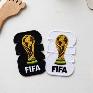 May include: Two embroidered patches featuring the FIFA World Cup trophy in gold, brown, and green. One patch has a black background with white text, and the other has a white background with black text. Both patches display the number 2022.
