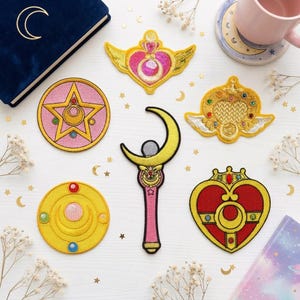 May include: A collection of six embroidered patches featuring various magical motifs including a pink star, a yellow crescent moon wand, a red heart, and golden winged emblems, all arranged on a white surface with scattered gold star confetti.