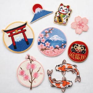 May include: A collection of embroidered fabric patches featuring Japanese-inspired designs including a red torii gate, Mount Fuji, a maneki-neko cat, pink cherry blossoms, a red Daruma doll with gold kanji, and two orange and white koi fish.
