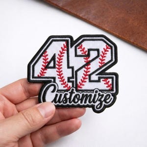 May include: A white baseball-themed patch with the number 42 in black outline, filled with red stitching, and the word "Customize" in a script font. The patch is held by a hand.
