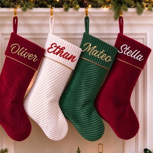 May include: Four ribbed Christmas stockings hanging in a row, featuring two deep red, one white, and one dark green stocking. Each stocking has a personalised name embroidered in cursive script: Oliver, Ethan, Mateo, and Stella.