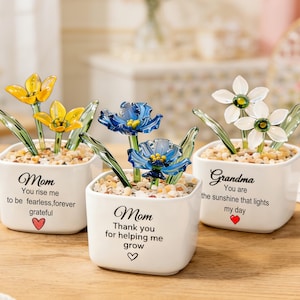 Könnte beinhalten: Drei kleine quadratische Keramiktöpfe in Weiß mit Glasskulpturen von Blumen. Einer hat gelbe Blumen, einer blaue und einer weiße. Jeder Topf hat den Text "Mom" oder "Grandma" mit einer Botschaft und einem Herzsymbol.