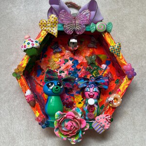 May include: A colorful, handmade wall hanging with a diamond-shaped frame. It features two wooden dolls, butterflies, hearts, and sweets. The frame is painted with vibrant colors and adorned with decorative elements, creating a whimsical and playful aesthetic.