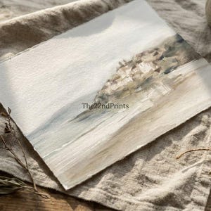 Puede incluir: Pintura a la acuarela de un paisaje costero con tonos suaves. La obra presenta una paleta de colores azul claro y beige, con una superficie de papel texturizado. El texto "The22ndPrints" es visible. La pintura se exhibe sobre un pa&ntilde;o de lino.