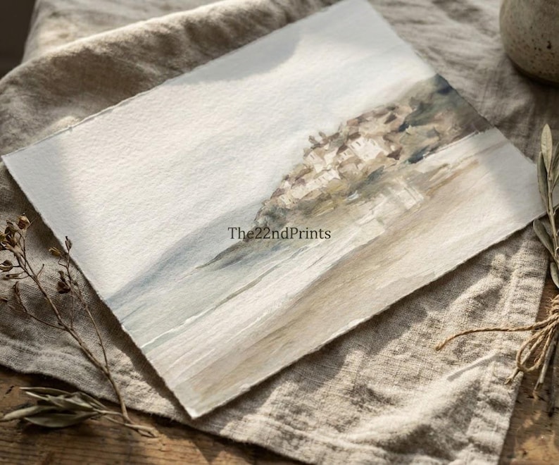 Puede incluir: Pintura a la acuarela de un paisaje costero con tonos suaves. La obra presenta una paleta de colores azul claro y beige, con una superficie de papel texturizado. El texto "The22ndPrints" es visible. La pintura se exhibe sobre un pa&ntilde;o de lino.