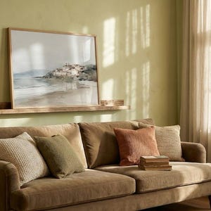 Puede incluir: Sal&oacute;n con un sof&aacute; marr&oacute;n y cojines en tonos beige, verde y naranja. Un cuadro enmarcado con un paisaje cuelga sobre el sof&aacute;, sobre una estanter&iacute;a de madera. La habitaci&oacute;n tiene una paleta de colores neutros.